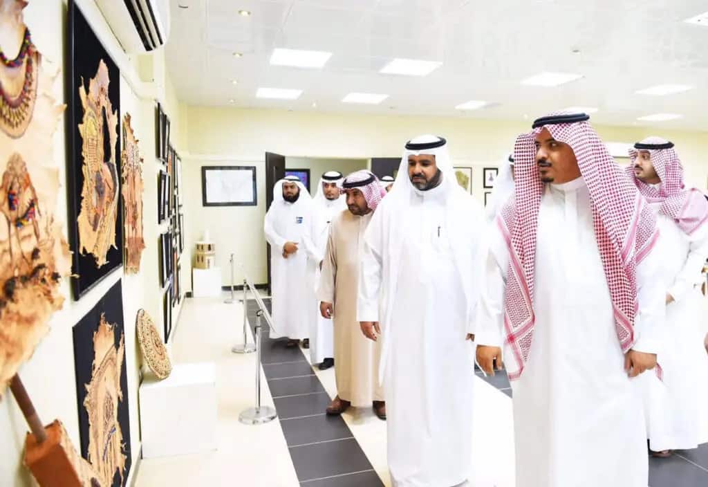 مجموعة من الرجال يرتدون الزي السعودي التقليدي - بما في ذلك الغترة أو الشماغ السعودي، والعقال، والبشت السعودي - يراقبون القطع الفنية المعروضة على جدران المعرض خلال معرض فن، الذي ينظمه كلية التصاميم والأسواق المنزلية في القصيم. يعكس هذا الواقع روح رؤية 2030 في قاعة عرض مضاءة بإتقان ومهيأة لاستقبال الزوار.