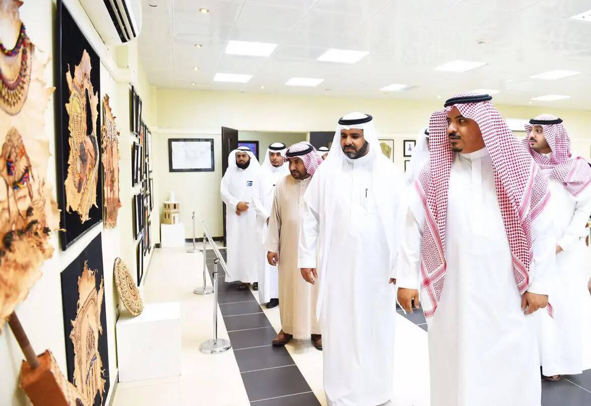 مجموعة من الرجال يرتدون الزي السعودي التقليدي - بما في ذلك الغترة أو الشماغ السعودي، والعقال، والبشت السعودي - يراقبون القطع الفنية المعروضة على جدران المعرض خلال معرض فن، الذي ينظمه كلية التصاميم والأسواق المنزلية في القصيم. يعكس هذا الواقع روح رؤية 2030 في قاعة عرض مضاءة بإتقان ومهيأة لاستقبال الزوار.