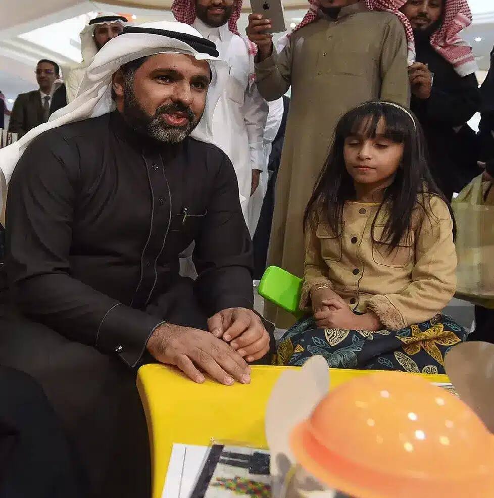 يجلس رجل يرتدي الغترة السعودية مع الشماغ والعقال والبشت السعودي بجوار فتاة صغيرة على الطاولة الصفراء، وذلك من خلال حملة توعوية في كلية التربية في القصيم، وسط حضور عدد من الناس. يبدو الرجل منتبهاً أثناء تحسين ملامح التفكير في الفتاة في هذا المكان الداخلي العام.