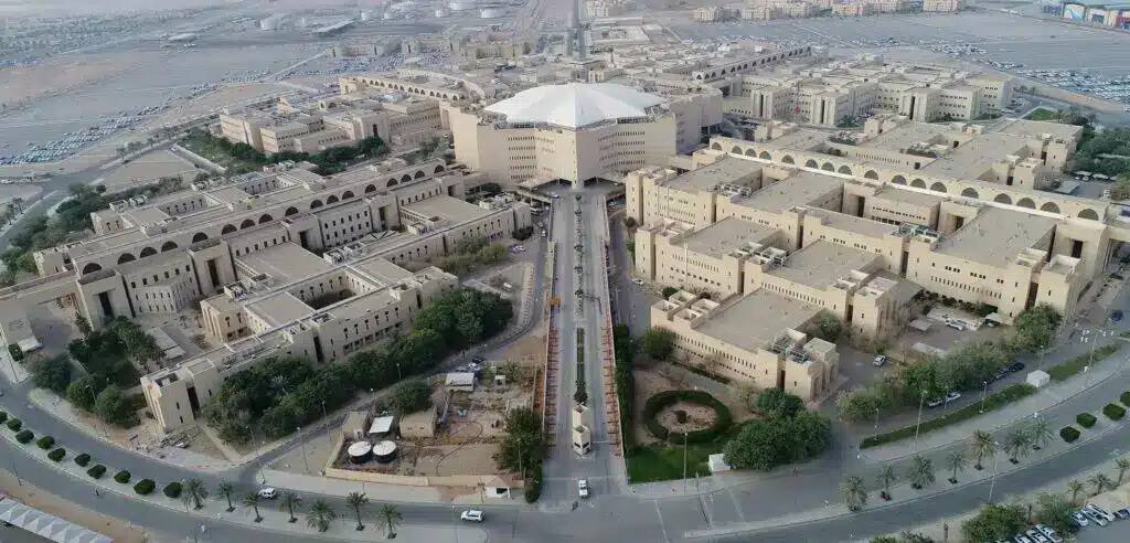 Aerial photo of Al-Qassim University Campus The modern campus with its buildings where the overall color is present, with a main building with a white roof, surrounded by tree-shaded roads and parking areas specially equipped for female students' cars. The overall landscape is upscale and commensurate with the nuclear nuclear environment, and students and staff stand out by wearing the Saudi shemagh or ghutra with the Saudi aqal and bisht at official events, emphasizing the national affirmation and cultural identity of the Kingdom of Saudi Arabia.