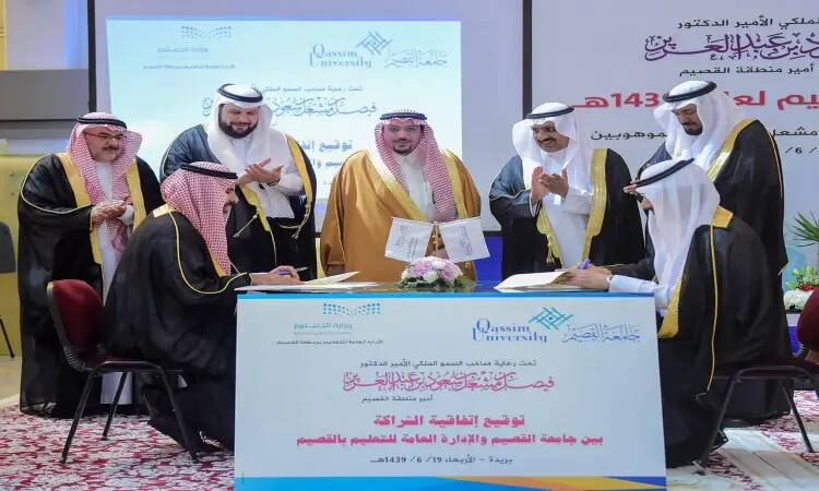 A group of men in Saudi Arabia's Ghutra, Shamaj, Egal and Bisht gathered around a signing ceremony table marking the beginning of a new partnership, where two of them signed documents in the presence of a crowd of smiling attendees. The table was surrounded by the banners of Qassim University, which were clearly visible in the background, adding another purpose and tracing the location of the university.