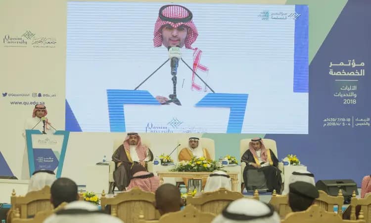 An adult dressed in a Saudi Arabian ghutra, headband and bisht, addresses attendees at a privacy conference in Qassim, while three men sit behind him on a podium. It contains full images on the main display screen, with text in Arabic appearing in Privacy Considerations and Challenges.