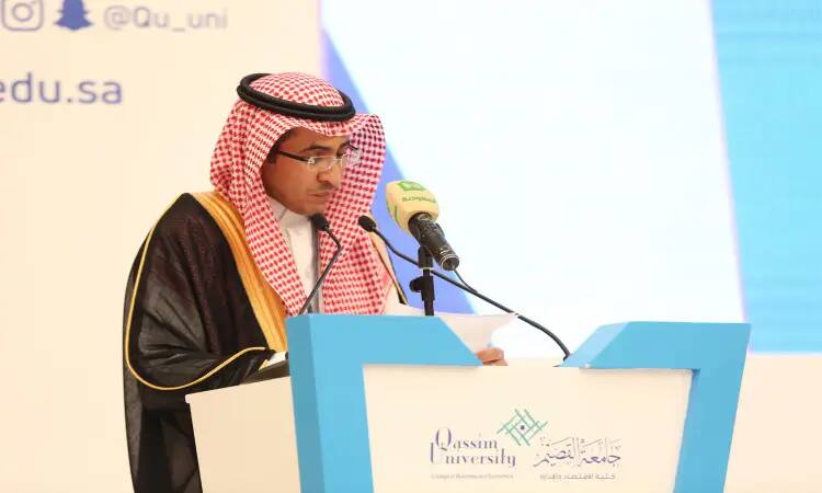 A man wearing a Saudi ghutra, shemaghqal and bisht behind a public speaking platform bearing the logo of Qassim University, writing with his hand. In front of him is a microphone with a green hood, while he picks up background texts in Arabic and English.