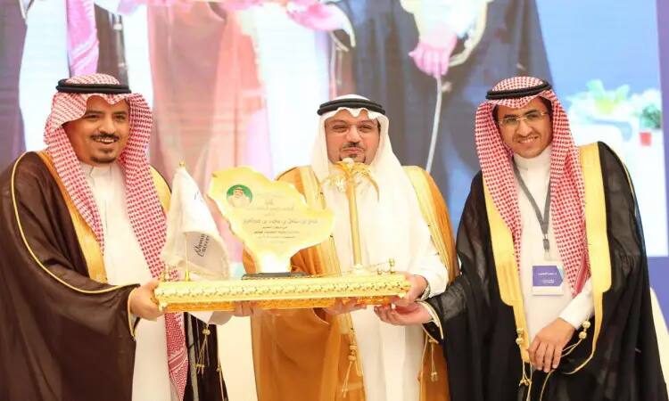 Contact Three men under the Saudi Ghutra or Shemagh, wearing the Saudi Eqal and Bisht, side by side, smiling as they hold a golden shield in reasonable honor during a privacy conference in Qassim. Behind them is a brightly lit backdrop and an unexplained photo in Paris.