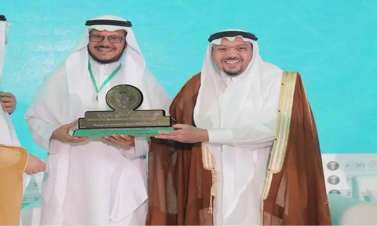 Two men dressed in the official Saudi Arabian ghutra and headband on a stage in the city of Buraida, smiling as they pose for a large plaque of honor. The logo of Qassim University and a blue background can be seen in the scene behind them. One of the two men stands out for investing in the colorful masht, highlighting agricultural opportunities.