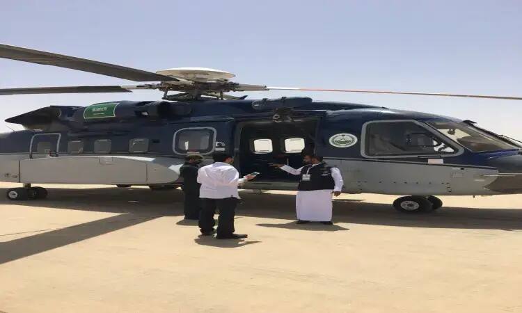 Three men called in to fly from a blue, covered blue airplane parked on a concrete deck under a clear sky. Two of them are dressed in a Saudi ghutra with a Saudi headband and bisht, while one appears to be preparing to enter military equipment, while the other two are chatting, presumably part of the team in Qassim.
