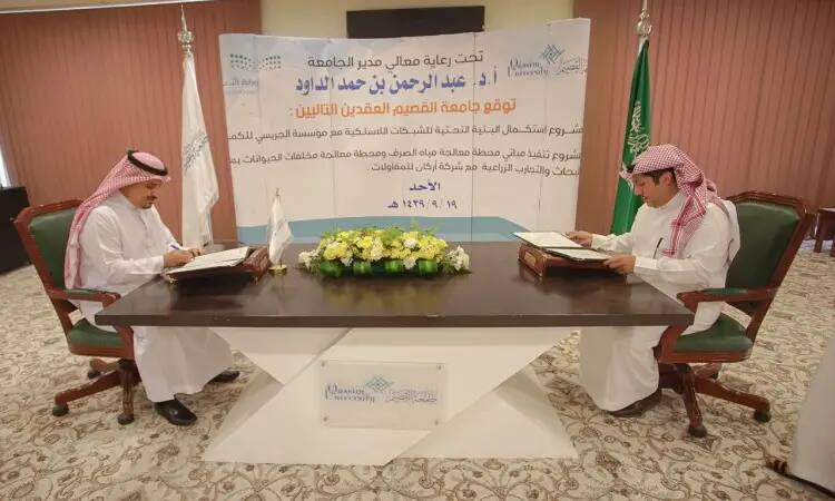 يجلس رجلان ويرتدي الغترة السعودية والشماع والعقال والبشت السعودي حول طاولة، يوقعتان وثائق تتعلق بمحطة المياه. احتياط خلفهما لافتة علمية وعلم المملكة العربية السعودية، مما يؤكد الطابع الرسمي لهذا الحدث التوقيع الرسمي في جامعة القصيم.