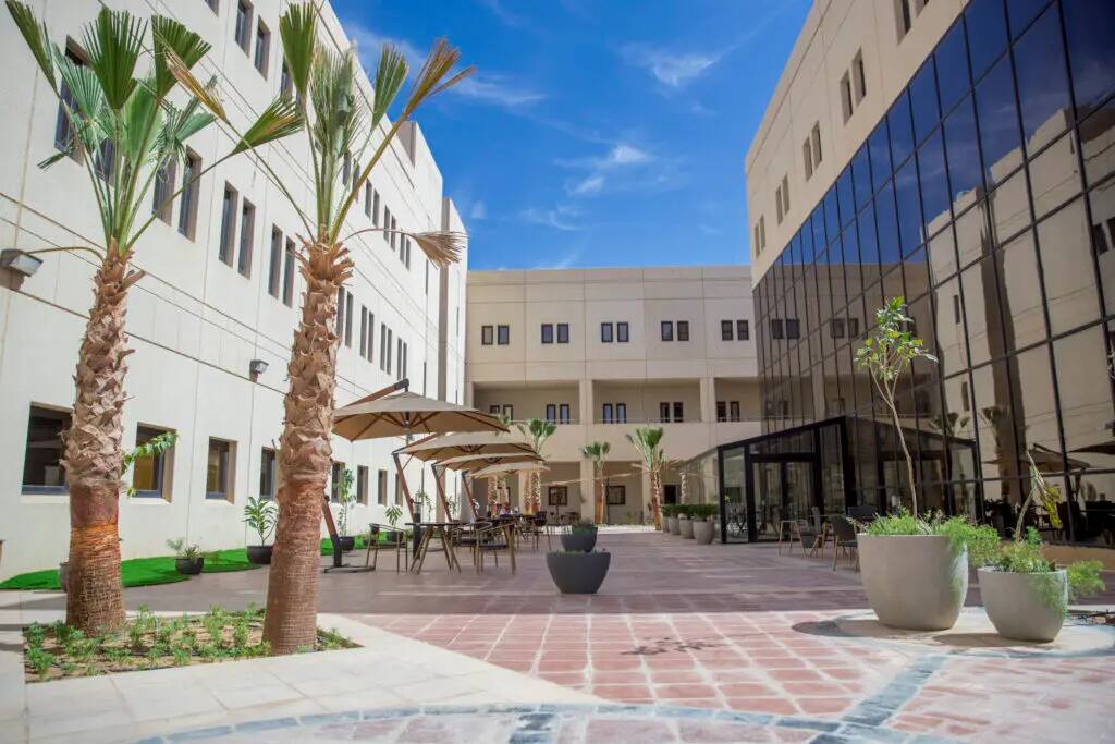ساحة تقع بين مبنيين جناحين ظللة في جامعة القصيم، تتزين بأشجار النخيل ونباتات كبيرة في أصص، طاولات وكراسيات خارجية مظللة بالمظلات، بينما تؤمن النوافذ الزجاجية وتدعم زرقة السماء. ندعوكم لتعلم جامعة القصيم.