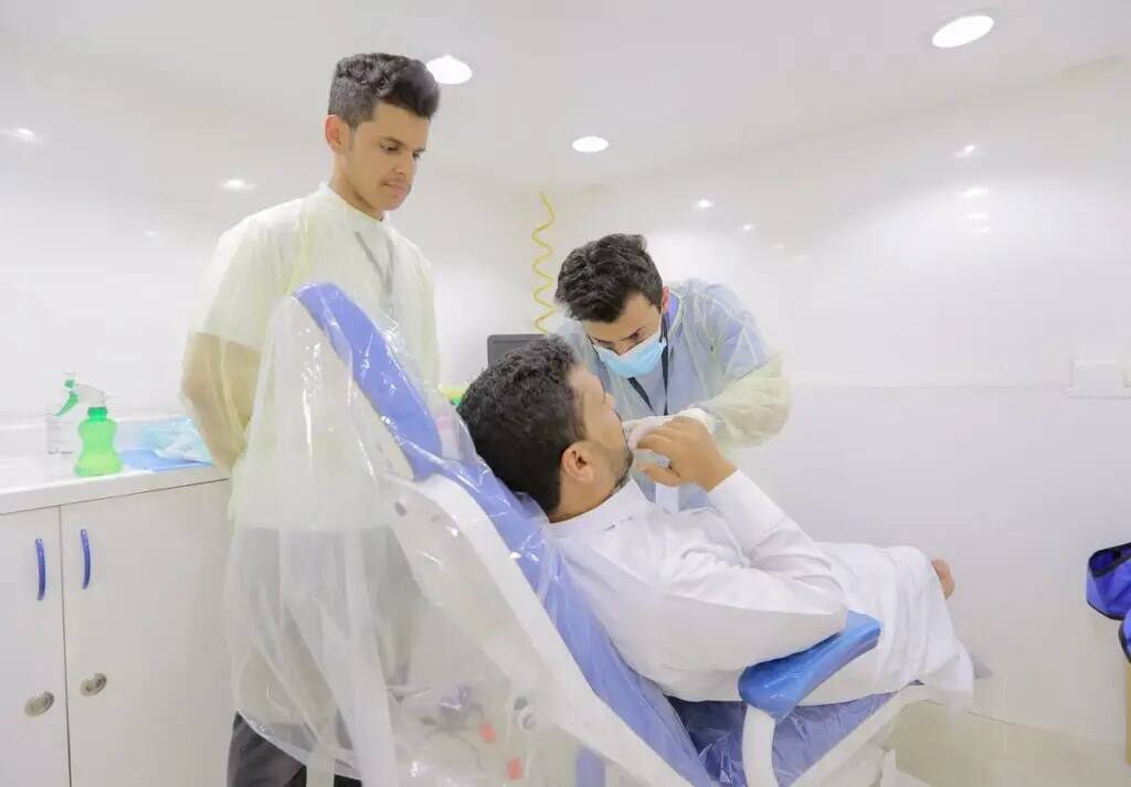 A doctor places a patient in a dental chair, while a dentist in Qassim Medical City meticulously checks his equipment. Standing next to him is another person dressed in a Saudi medical coat and shemagh, observing the filing procedures in the clean and brightly lit clinic room.