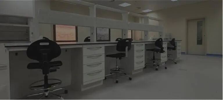 A modern laboratory is equipped with high standards for nursing education, as it includes white work units with precise organization, and black mobile chairs equipped with wheels to facilitate movement. The laboratory contains drawers and partitions between stations, under bright ceiling lighting that ensures visibility, and the laboratory is characterized by complete cleanliness and order, and there are windows on the far side that allow medical light to enter, in addition to a closed door located on the right side of the place.