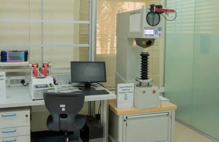 تضمين محطة العمل المخبرية المخصصة لتخصص الهندسة الميكانيكية في جامعة القصيم جهاز جهاز حاسب حديث، وكرسي أسود، وجهاز اختبار الصلادة الشامل من نوع من زايس (جهاز اختبار الصلابة العالمي من زايس)، بالإضافة إلى معدات مخبرية أخرى موضوعة على طاولة قريبة من النوافذ الزجاجية ذات الصلابة الستائر.