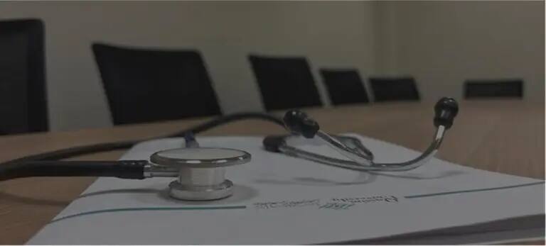 يرتفع سماعة الطبيب فوق مستند موضوع على طاولة اجتماعات خشبية، في مشهد يرمز إلى صحة المجتمع وتظهر في الخلفية كراساً سوداء خالية، ويُضفي على ذلك كله طابعاً رسمياً يناسب جامعة القصيم.