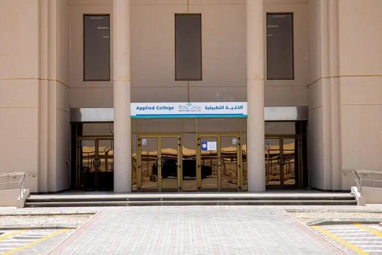 مدخل مبنى رمادي داكن بدون أبواب، تعلوه لوحة تحمل اسم "الكلية التطبيقية" باللغتين العربية والإنجليزية، ويستند المدخل إلى أربع أعمدة كبيرة. ويقود ممر مرصوف بالطوب إلى بوابة الدخول.