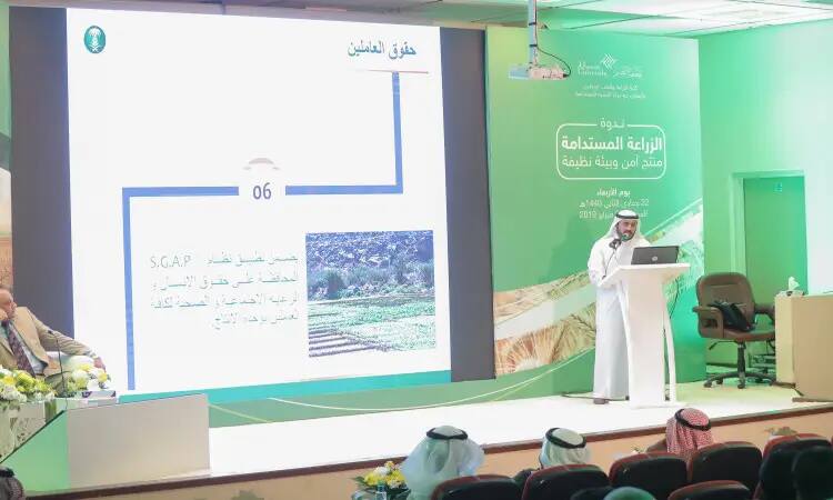 Dressed in a Saudi ghutra or shemagh with a Saudi aqal and besht, he gathers in front of the podium as he delivers his speech at a symposium on agriculture sponsored by Qassim University. A screen behind him displays Arabic text and an image of a farm, highlighting the theme of sustainability in the agricultural sector, while the audience watches intently.