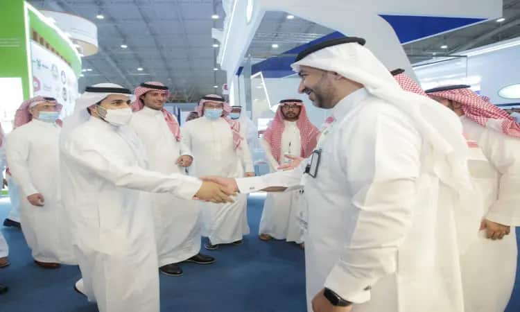 A number of men wearing the Saudi Ghutra or Shemagh, along with the Saudi Aqal and Bisht, gather in a house in Riyadh, some wearing masks. They exchange greetings and shake hands in the center of the room, keeping behind them the pavilions and banners of the Middle East Poultry Exhibition.