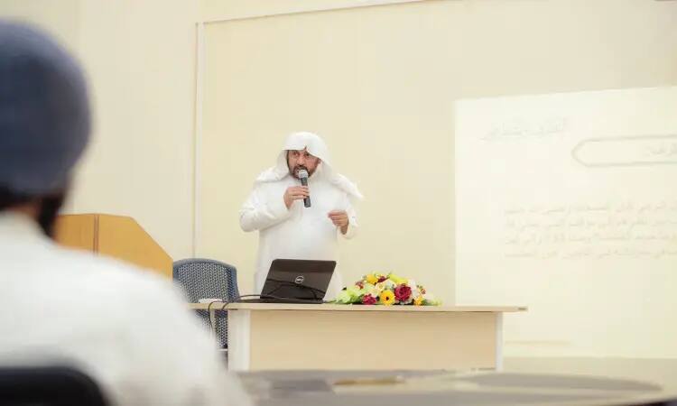 ويرتدي رجلٌ غترة سعودية وقالاً وبشتاً سعودياً، ويتحدث عبر مكبر الصوت إلى الحضور بجانب جهاز الكمبيوتر المحمول وباقة الزهور. وخلفيه شاشة عرضها نص باللغة العربية يتناول موضوع التحصين الفكري، وذلك في قاعة الحضور في القصيم.