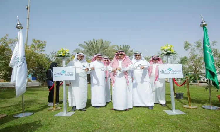 مجموعة من الرجال الحاضرين الغترة أو الشماغ السعودي مع العقال والبشتاء السعودي، الاتصالون في الهواء الطلق على المسطحات الخضراء خلال مراسم لزراعة الأشجار في جامعة القصيم. يظهرون شريط مبتسمين وهم يبدأون الافتتاح، ويحيط بهم عمودان للعلم مزينان للانضمامات ولا الزهور على الروابط، بينما تبدو الأعمال والسماء الصافية في.