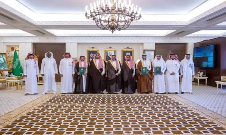 Contact A group of men in their traditional Saudi attire, wearing the Ghutra or Shemagh with the Saudi Eqal and Bisht, with His Highness the Emir of Qassim Province in the center, inside the men's hall. An era with only new elements that have nothing to do with the anniversary of understanding. In the background is the flag of the Kingdom of Saudi Arabia and a portrait of its leadership, while a chandelier hangs from a panel out of respect for the official venue.