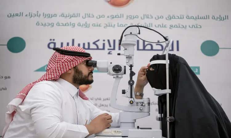 An official optometrist wearing the Saudi Arabian ghutra, aqal and bashta examines the eyes of women in black using a slit, as part of eye health care and awareness within the clinic at Qassim University facilities, with Arabic text in the background that properly expresses the university's mission and interests.