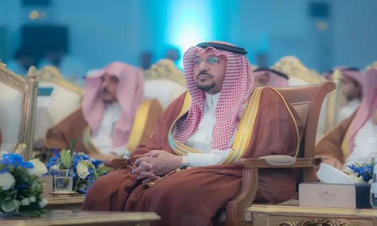 A man dressed in a Saudi bisht, wearing a white shemagh and wearing a fourth headband, sits in a plush chair during the University of Qassim's mid-year convocation in the presence of His Highness the Emir of Qassim. Adjacent tables adorn the green format at the official honor gate.