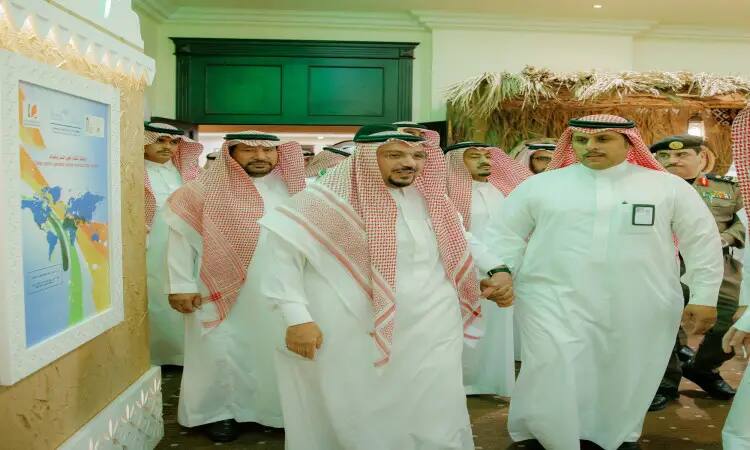 A group of men in traditional Saudi attire, the white Saudi Ghutrah with the red shemagh and aqal, as well as the Saudi besht. They walk inside the university exhibition during the Buraydah Dates Carnival, next to a decorated wall. A colorful structure and a structure made of palm fronds are also visible, adding to the heritage of the event hosted by Qassim University.