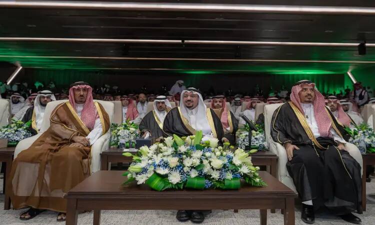 A group of men wearing the Saudi ghutra or shemagh with the Saudi aqal and bisht, led by His Highness the Emir of the region, in the front row of one of the official National Day events organized by Qassim University. The presence of the zahriyah gives the tables a distinctive formal look, as the audience in the hall behind them is crowded with a wide participation of university staff and invitees.