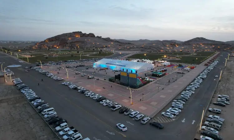 An aerial view of the fifth building construction event at the Mudkhana Center, where the university attendees are seen with a number of participants wearing the Saudi Ghutra, Aqal and Bisht, as a distinctive feature includes a tent structure illuminated with decorative lights to rest cars parked at the foot of the mountain alone at sunset, all within an awareness and education framework that reflects the official and organizational character of Qassim University.