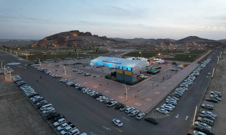 An aerial view of a major event organized by Qassim University in a brightly lit tent, dedicated to the Pillow Awareness Forum; wearing cars and compensatory lanes in the heart of the desert, replaced by rocky hills with a sunset backdrop. Attendees gather in their formal attire, wearing the Saudi Shemagh or Ghutra and Eqal, as well as the Saudi Bisht, which lends the event a sense of dignity and formality.