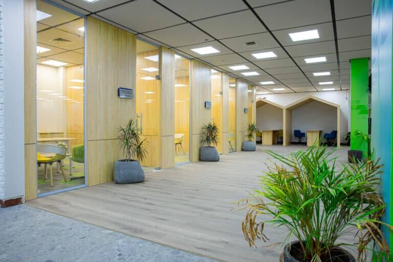 A modern office space that includes meeting rooms with glass tables on the left side, coordinated plants distributed along the corridor in addition to a corner for individual work with chairs on the right side; this environment is ideal for information systems teams or production management within an open and light space with integrated ceiling lighting. The design takes into account the formal and practical design suitable for Qassim University with the possibility of emphasizing the national identity by wearing the Saudi Shemagh or Saudi Ghutrah according to the customs and traditions followed at the university.