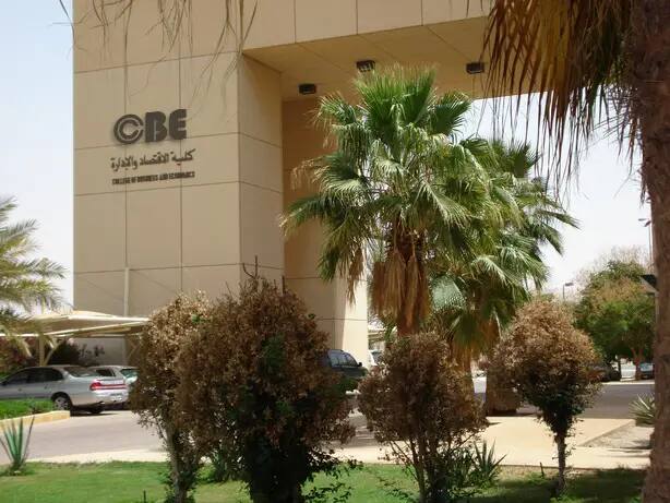 A modern building bearing the sign "College of Economics and Administration" "cbe" in Arabic and English, surrounded by a vegetable carpenter and vegetable trees, with the Department of Information Systems among its departments. Cars parked near the building on a sunny day, as part of the official environment of Qassim University.