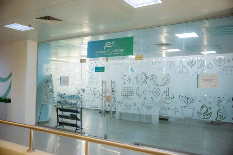 A glass-walled office decorated with eco-themed graphics and Arabic phrases, including the words "Center for Sustainable Development" written on the glass, a file rack inside the office, and a bright ceiling light that illuminates the space.