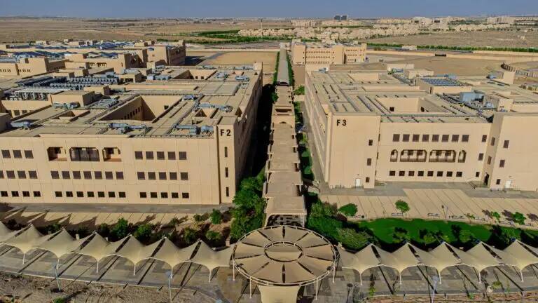 صور جوية لمبيني لمبيني جامعيين كبيرين بلونين بلونين بيجون في جامعة القصيم هما كلية التصاميم (و2) والمبنى (و3)، يفصل بينهما ممر يتوسطه مظلات هندسية التصميم، وتحيط بهما مساحات خضراء ضمن بيئة صحراوية.