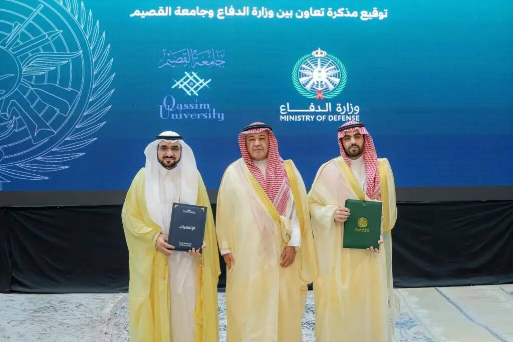 Three men in authentic Saudi attire, wearing the Saudi shemagh or ghutra, holding documents titled with a draft license, in front of a blue background with the logos of Qassim University and the Ministry of Defense, signifying the signing and cooperation between the two entities.