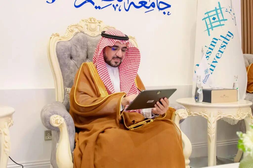 يرتدي الرجل الزي السعودي التقليدي مع شماغ سعودي على كرسي فاخر، ويطالع جهازاً لوحياً لاستخلاص مسعودة تلقائية. لاتخاذ راية كتب عنها "جامعة القصيم"، إلى جانب لمزيد من المعلومات عربي مزخرف على الحائط.