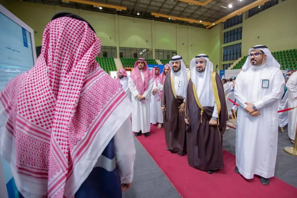 A group of men dressed in traditional Saudi dress, including the shemagh or ghutra in Saudi Arabia, walk across a red carpet in a large hall, where they find another man in formal attire facing them with his back to the camera, in a scene that emphasizes the first goal during an important meeting or introduction at Al-Qassim University.