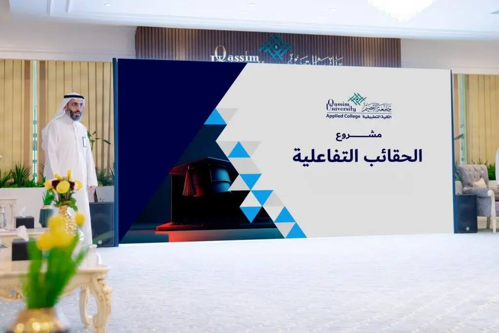 رجل يرتدي الزي السعودي الرسمي مع شماغ أو غترة سعودية شاشة كبيرة تظهر نصاً باللغة العربية وشعار الكلية التطبيقية، وذلك في قاعة الزينة الحديثة بالنباتات والزهور الزخرفية، حيث يدخل الحقائب التفاعلية.