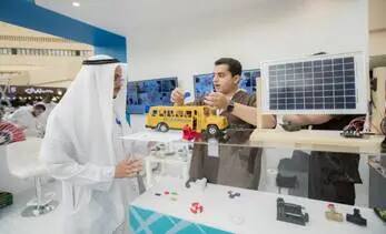 يرتدي رجل يرتدي شماغ السعودي أثناء استماعه لشرح حضوره أحد المشاركين حول نموذج لعبة أصفر تعمل بالطاقة الشمسية من خلال لوح صغير، وذلك في قاعة الفعاليات الفنية، في تجسيدٍ للابتكار الذي يدعم الهدف الخامس عشر.