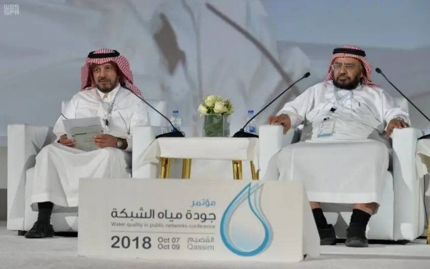 Two men wear the Saudi shemagh on stage, exchanging homely clothes next to a table adorned with flowers and water bottles. They are attending the Water Quality in Public Networks Conference, the sixth goal of the Sustainable Development Goals, taking place October 7-9, 2018, in Qassim.