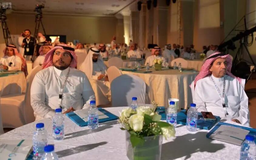 A group of men dressed in Saudi Arabian shemagh or ghutra and traditional costumes sit in an indoor hall, where tables are dotted with water, flowers and a few documents, as they attend an official conference discussing a report related to Goal 6. Some are listening intently to the discussion, while Akra looks around the room.
