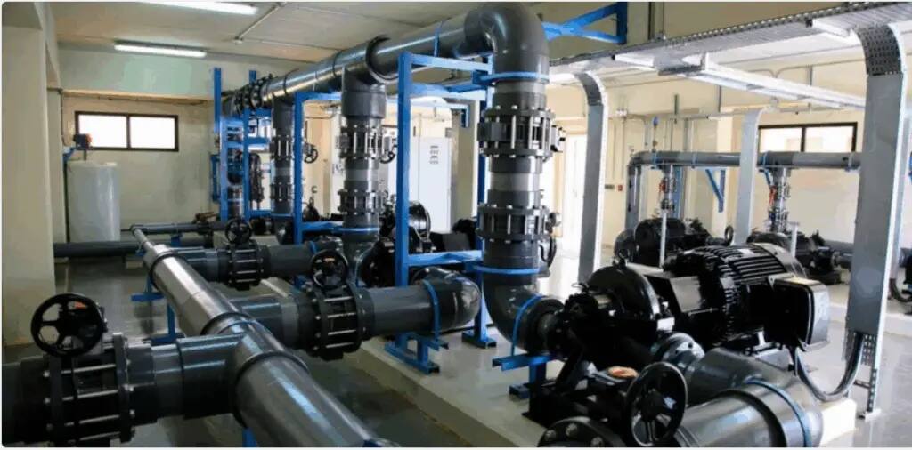 A clean and well-lit industrial room contains large black and gray pipes, blue metal structures, as well as valves, pumps, and machinery intended to treat water or liquids, in support of Goal 6, as stated in the report.