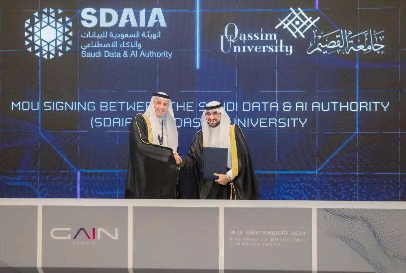 Two men wearing the Saudi shemagh and traditional dress exchange handshakes and smiles as they press a file stand in front of a sign bearing the logos of the Saudi Fraud and Artificial Intelligence Authority and Qassim University, at a signing event supporting Mr. Target IX.