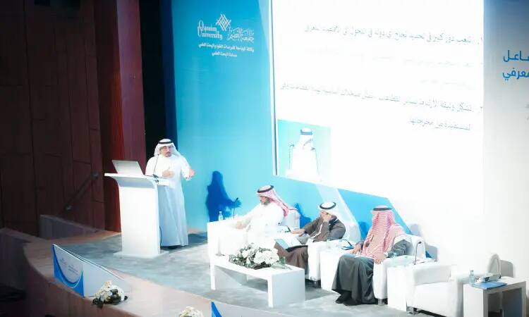 The speaker, dressed in traditional Saudi garb, including a Saudi shemagh, addresses the audience from the stage, while three men, equally well-dressed and wearing Saudi shemaghs, sit next to him in white chairs. A large rear projection screen carries text in Arabic and highlights the "ninth goal."