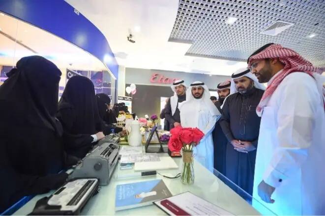A number of men, wearing the Saudi Shemagh or Ghutrah and traditional dress, gather around a table displaying a bouquet of flowers and a large number of books, reports and some equipment, in a modern environment. The atmosphere is formal and interactive, in line with the achievement of the tenth goal.