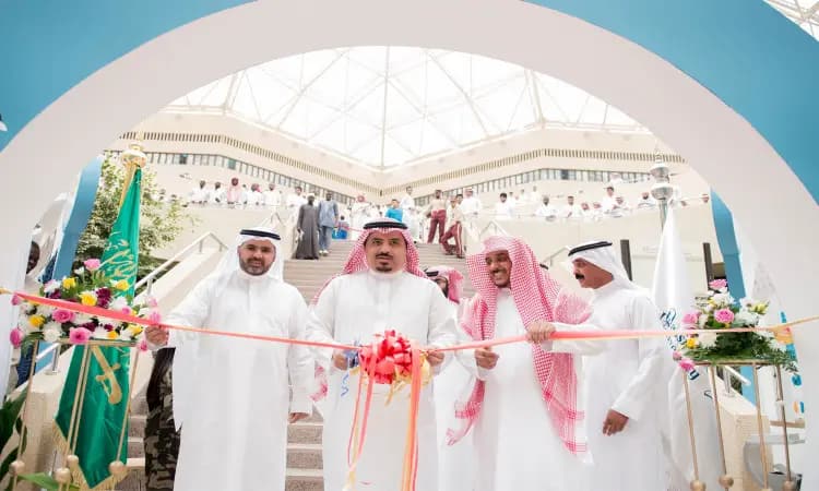 A group of men in authentic Saudi Arabian attire, including a shemagh or Saudi Arabian ghutra, cut the ribbon to mark the opening of an event at Qassim University, as part of achieving Goal 11. Write in background writing flags, flowers, and a crowd of moderns to connect to the degrees.