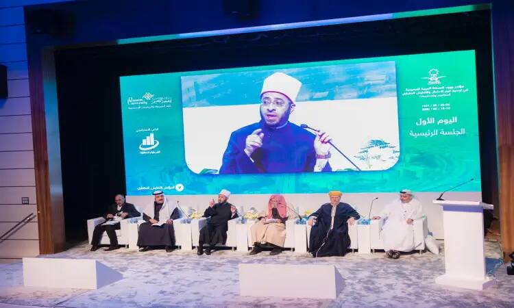 تلبيس ستة رجال الزي السعودي الأصلي، بما في ذلك الشماغ أو الغترة السعودية، على منصة رسمية ضمن حلقة نقاش خلال مؤتمر في القصيم. بوضوح وهو يشير بيده، بينما تُعرض صورته على شاشة كبيرة خلفهم بنصوص عربية وأبرز النقاط المستخلصة من تقرير الهدف الرابع.