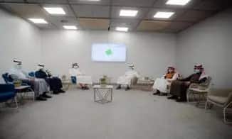 A group of six men in authentic Saudi dress, including the Saudi Shemagh or Ghutrah, are spaced apart on chairs in a modern, minimalist room, discussing a report on the 13th goal. A monitor behind them is mounted on the wall, with small tables and plants scattered among their chairs.
