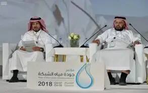 Two men dressed in traditional Saudi dress, including a shemagh or Saudi ghutra, stand on a plain podium with microphones and papers. It contains the main menu and bears Arabic text, a drawing of a drop of water and the year 2018. A different vase is placed on a fifth flower.