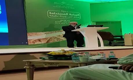 From a podium in a hall with a green backdrop and text in Arabic, a man spoke during a seminar on agriculture at Qassim University. Tables with drinks and snacks are set up at the front.