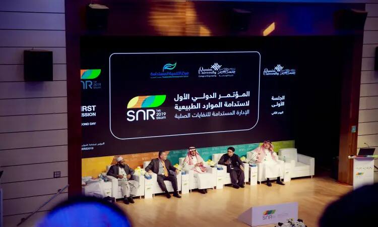يجلسون على المنصة خلال مؤتمر في جامعة القصيم، أمام شاشة كبيرة تعرض نصوصًا باللغة العربية والإنجليزية حول الفعالية الصعبة بالموارد الطبيعية، وتبرز على شاشة شعارات SNR 2019 ولافتات مع إشارات إلى "تقرير" و"الهدف الثاني عشر". أخذت بعض المشاركين الشماغ السعودي أو الغترة السعودية، بما في ذلك الطابع الرسمي للفعالية.