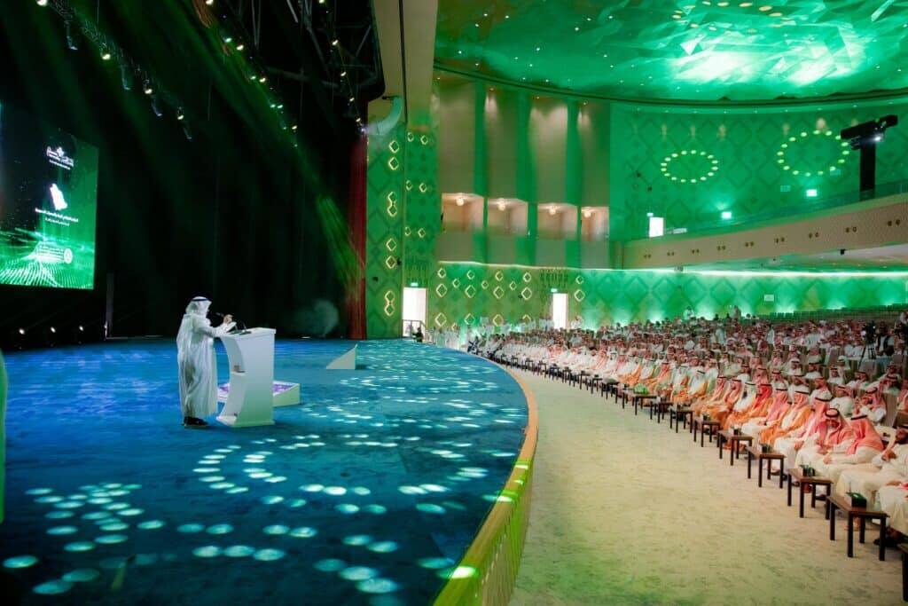 A lecturer delivers a speech at the Geography Conference at Qassim University's Haditha Hall with a large academic audience.
